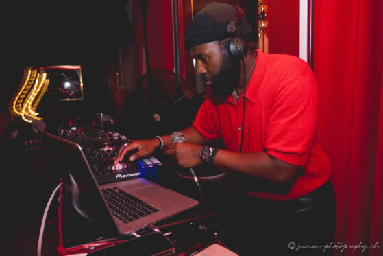 DJ Jakonet en pleine performance live à l’Élégancia Party 2025, mixant sur ses platines Pioneer, vêtu d’une chemise rouge, sous une lumière tamisée et dorée.